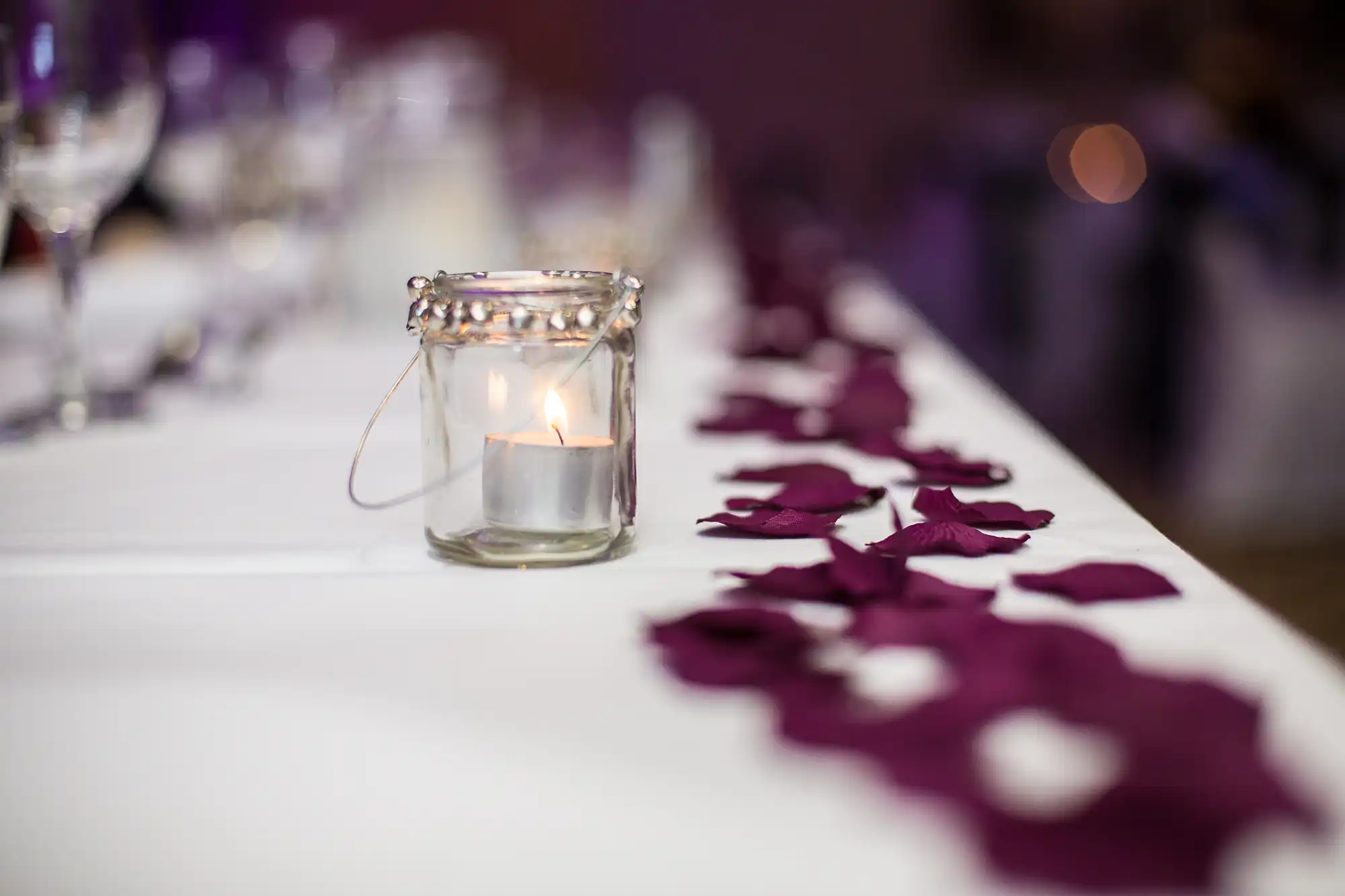 Glass candle holder with a lit candle on a banquet table decorated with purple petals, focusing on the foreground with a blurred background.