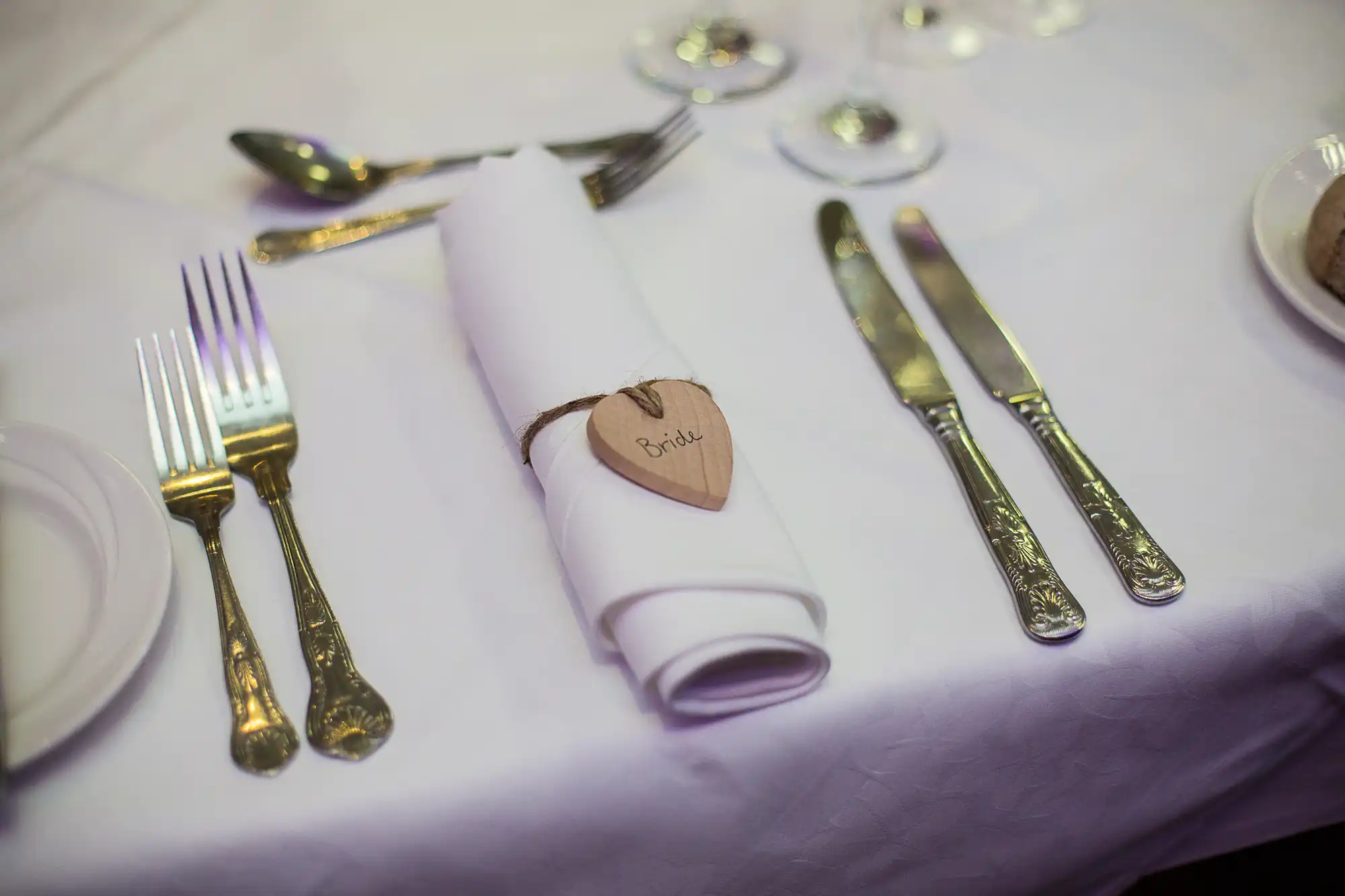 Formal dining setting with a rolled napkin labeled "bride," forks, knives, and glasses on a white tablecloth.