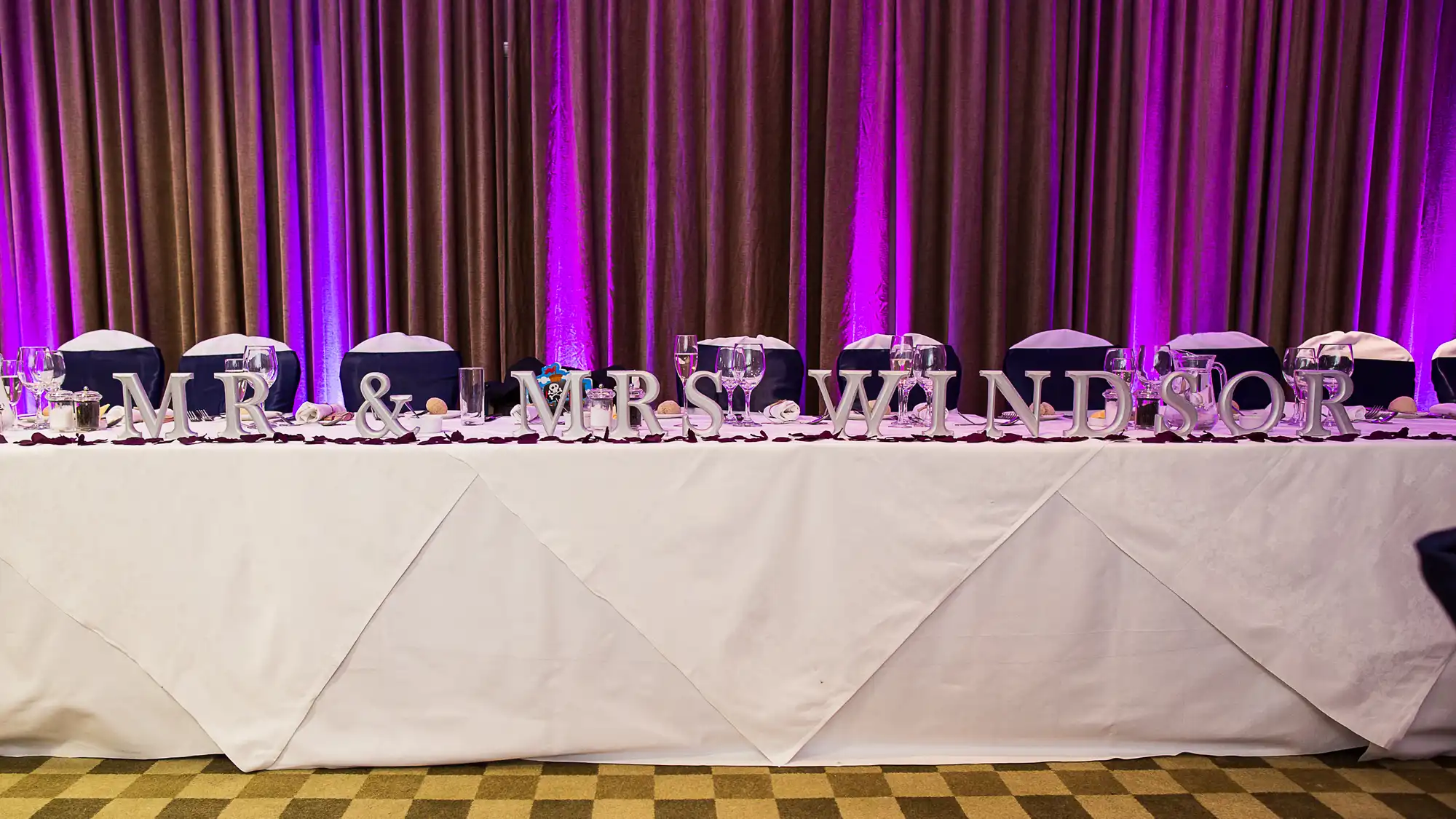 Decorated banquet table with "mr & mrs windhor" written in large white letters at a wedding reception, with purple lighting in the background.