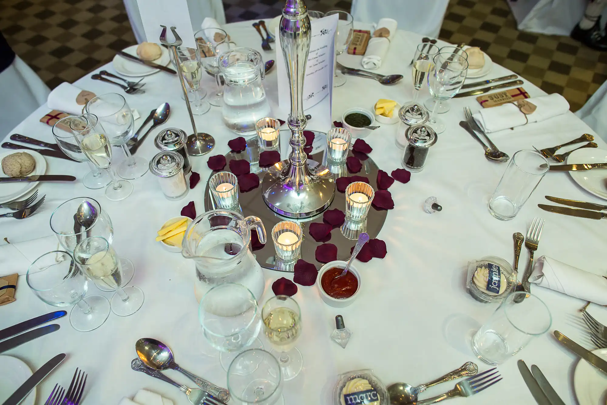 An elegantly set banquet table with silverware, glasses, candles, and scattered rose petals.