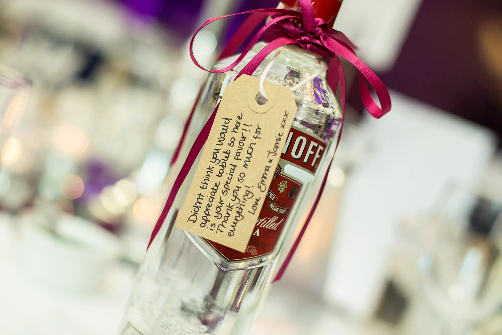 Close-up of a smirnoff vodka bottle with a custom handwritten label attached by a red ribbon, used as a wedding favor, on a festively decorated table.