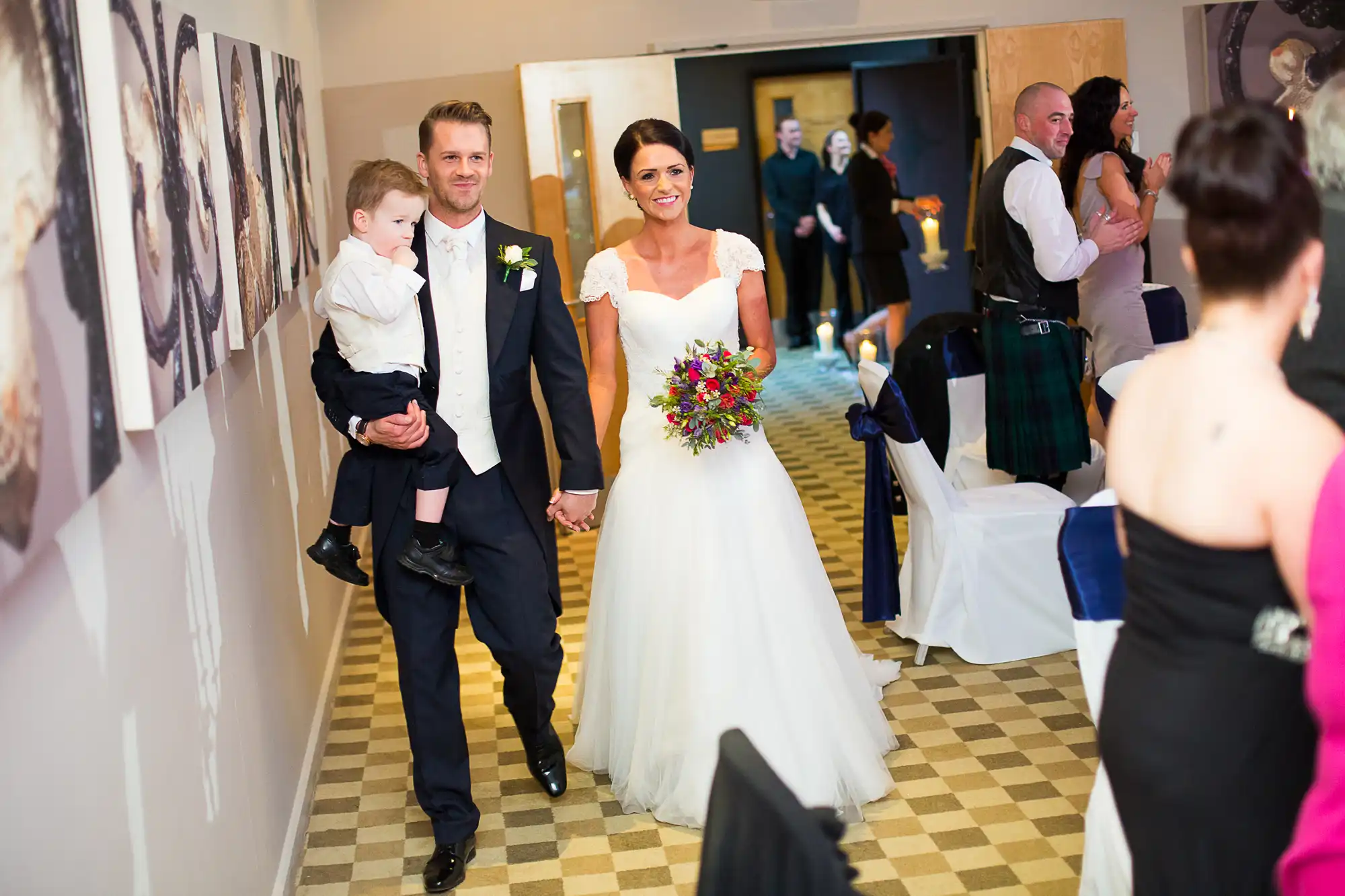 Bride and groom holding a young child, walking down a hallway lined with guests and framed pictures, smiling at the camera.