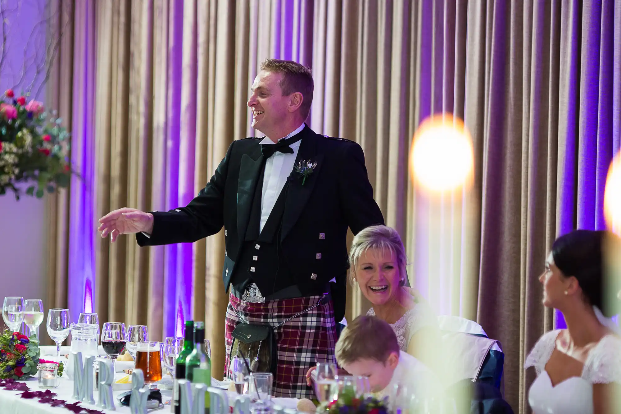 A man in a kilt and tuxedo jacket smiling and gesturing at a wedding reception, with seated guests laughing and enjoying around him.