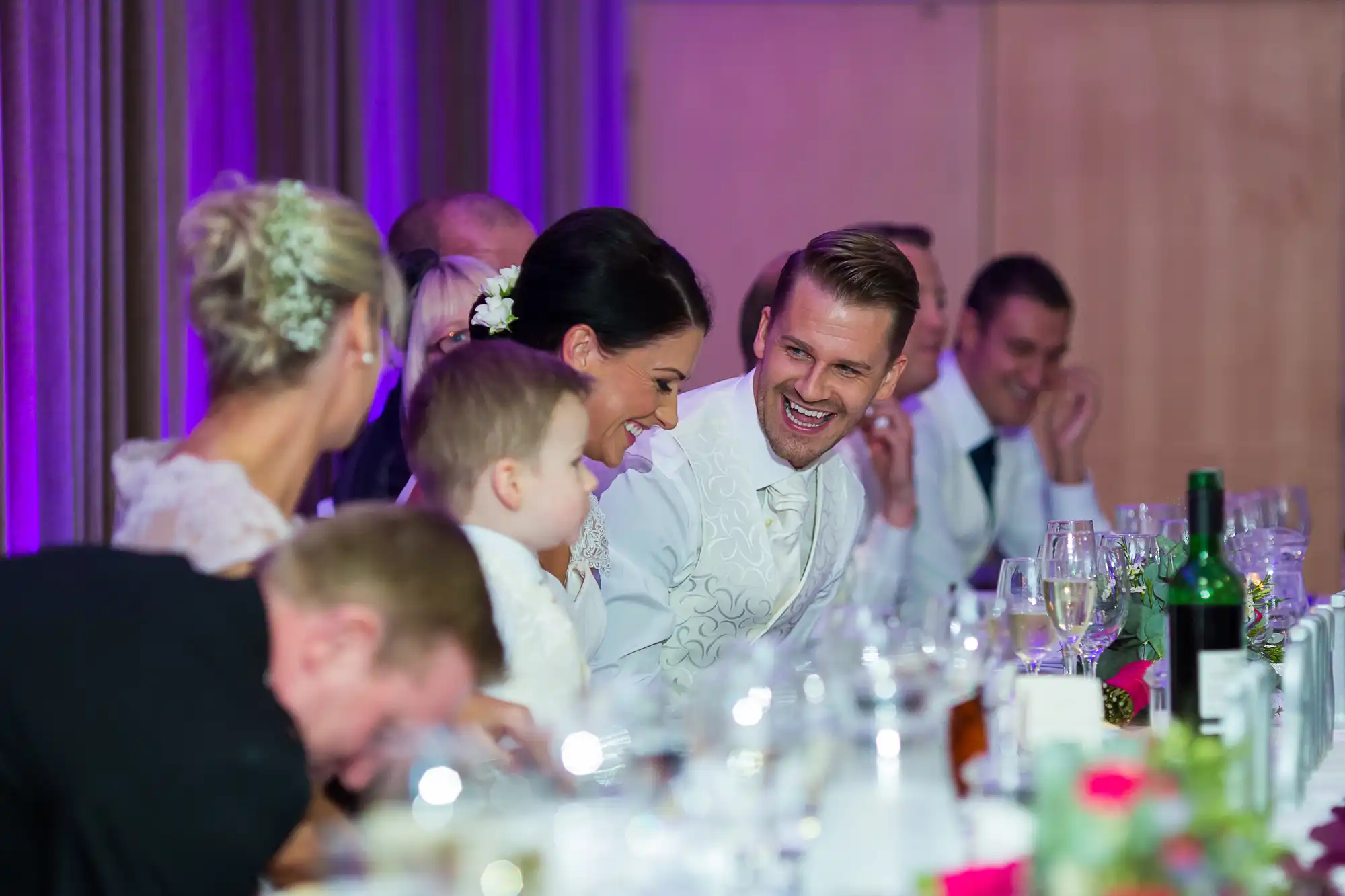 A joyful couple laughing at a wedding reception table surrounded by guests and decorated with elegant dinnerware.