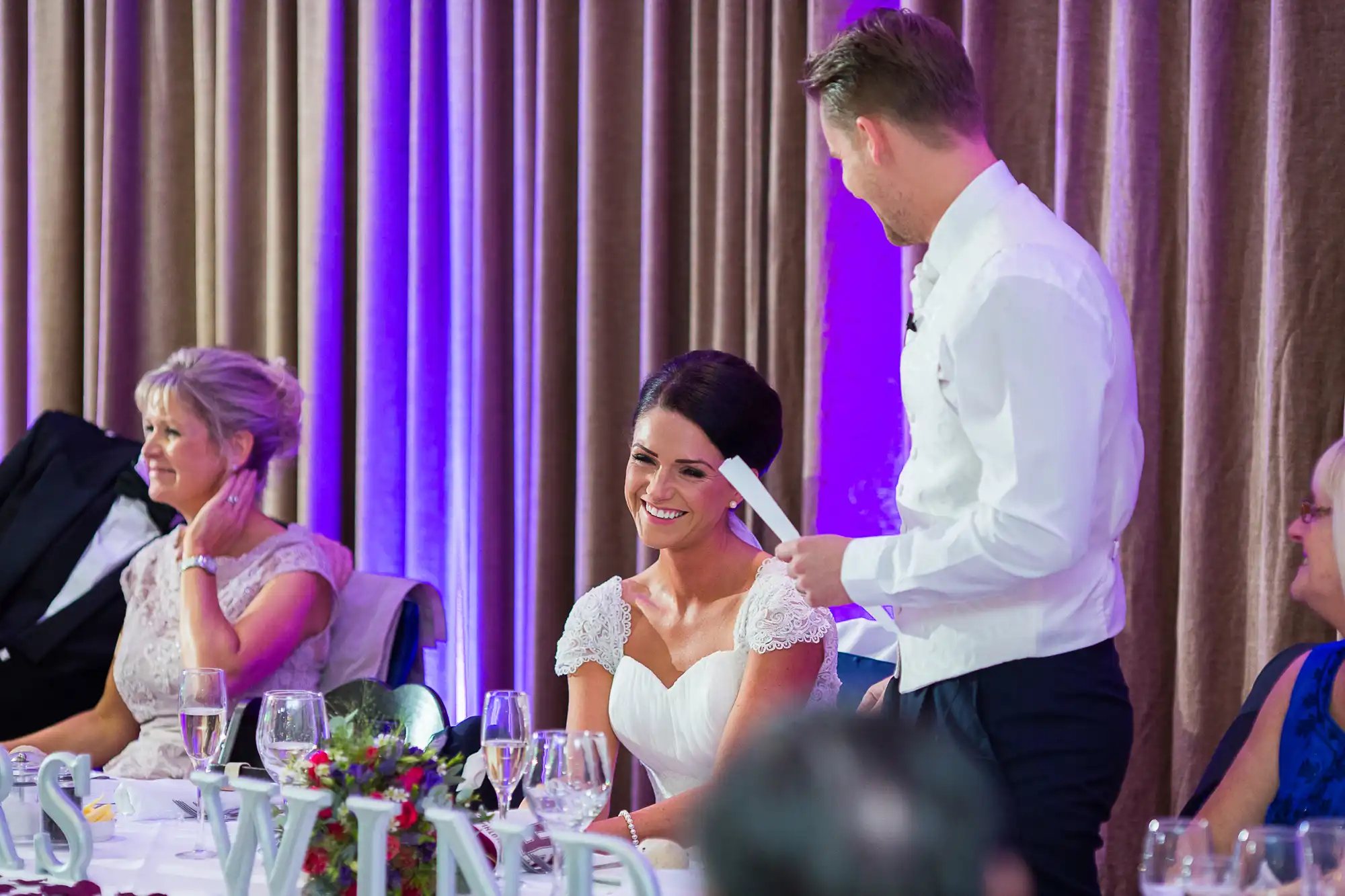 A smiling bride in a white dress seated at a wedding reception table, with a man standing beside her pointing playfully at her cheek.