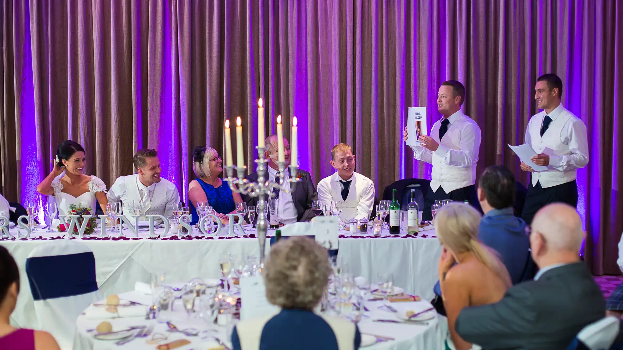 Two men standing and giving a speech at a wedding reception while guests seated around tables listen attentively.