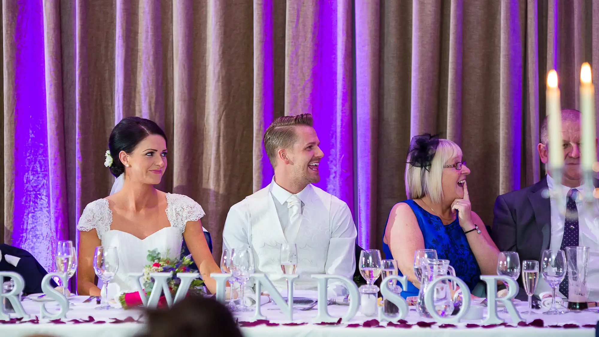 A bride and groom seated at a banquet table with a guest, all smiling, in a room with purple lighting and elegant decor.