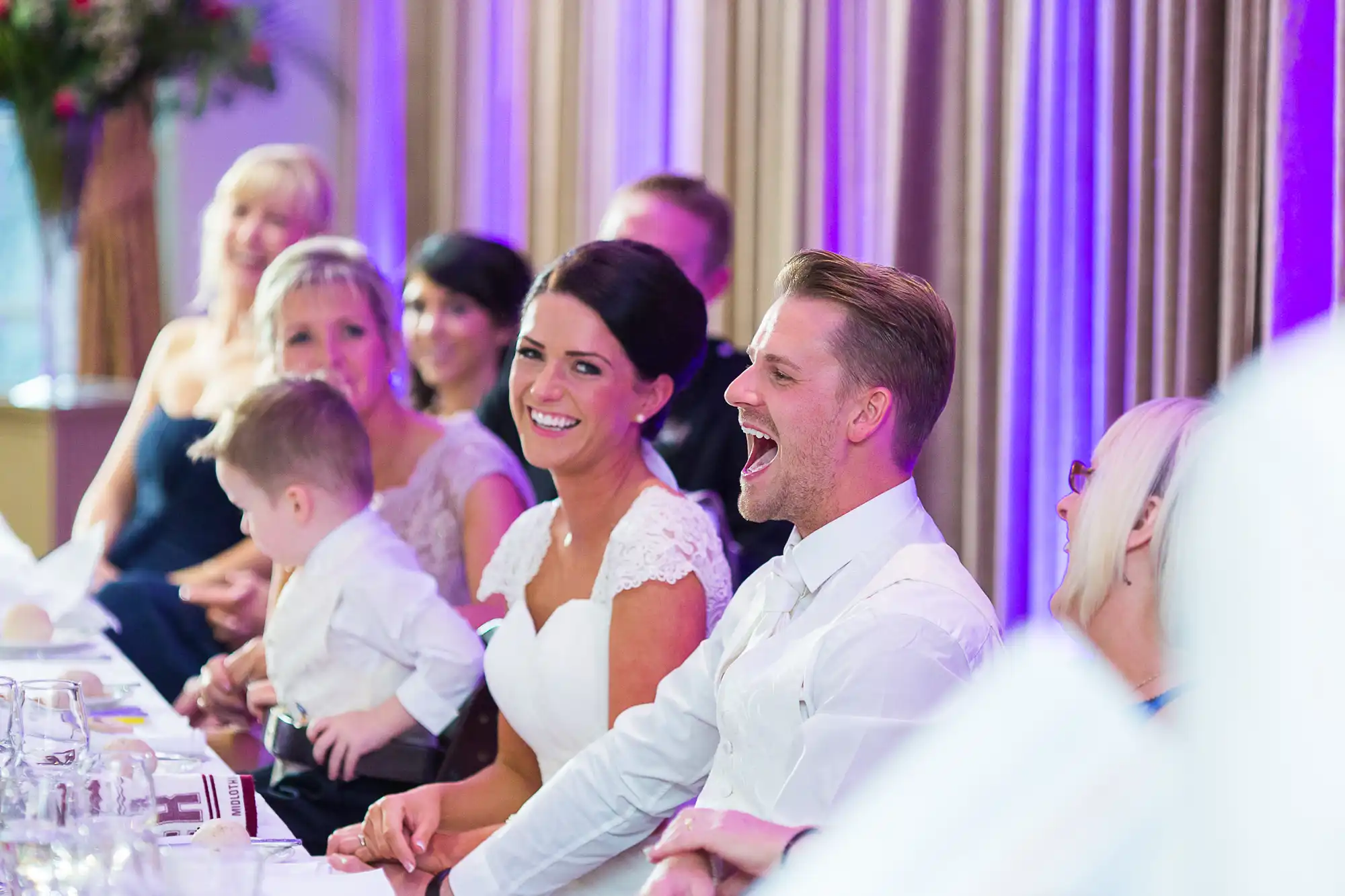 A bride and groom laughing joyously at a wedding reception table, surrounded by guests in a festively lit venue.