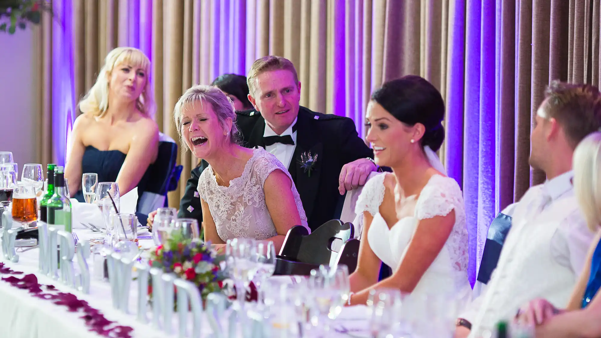 A wedding reception scene with guests sitting at a decorated table, a bride in white conversing with a woman laughing, and a man in a military uniform smiling behind them.