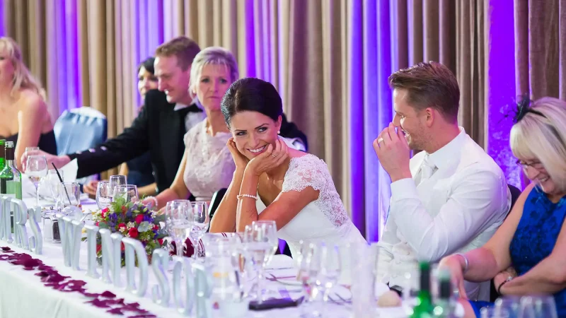 A bride seated at a wedding reception table laughs, resting her chin on her hand, while surrounded by guests in a festively decorated venue.