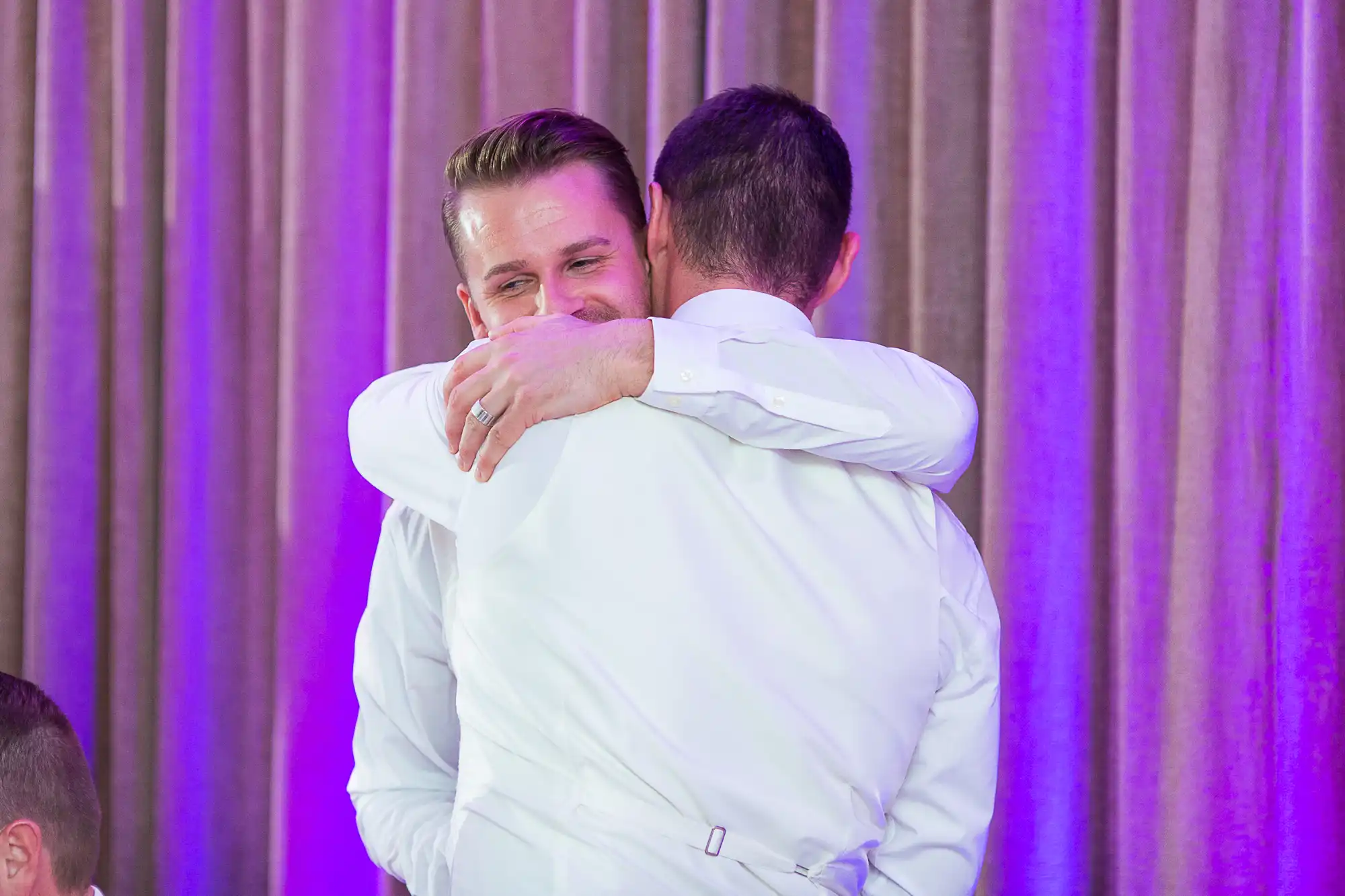 Two men in formal wear sharing a heartfelt embrace at an event with a purple-lit background.