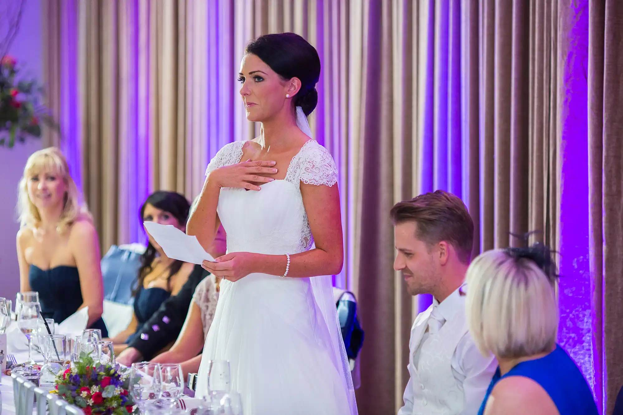 A bride in a white lace dress stands giving a speech at a reception, while guests at the dinner table watch attentively.
