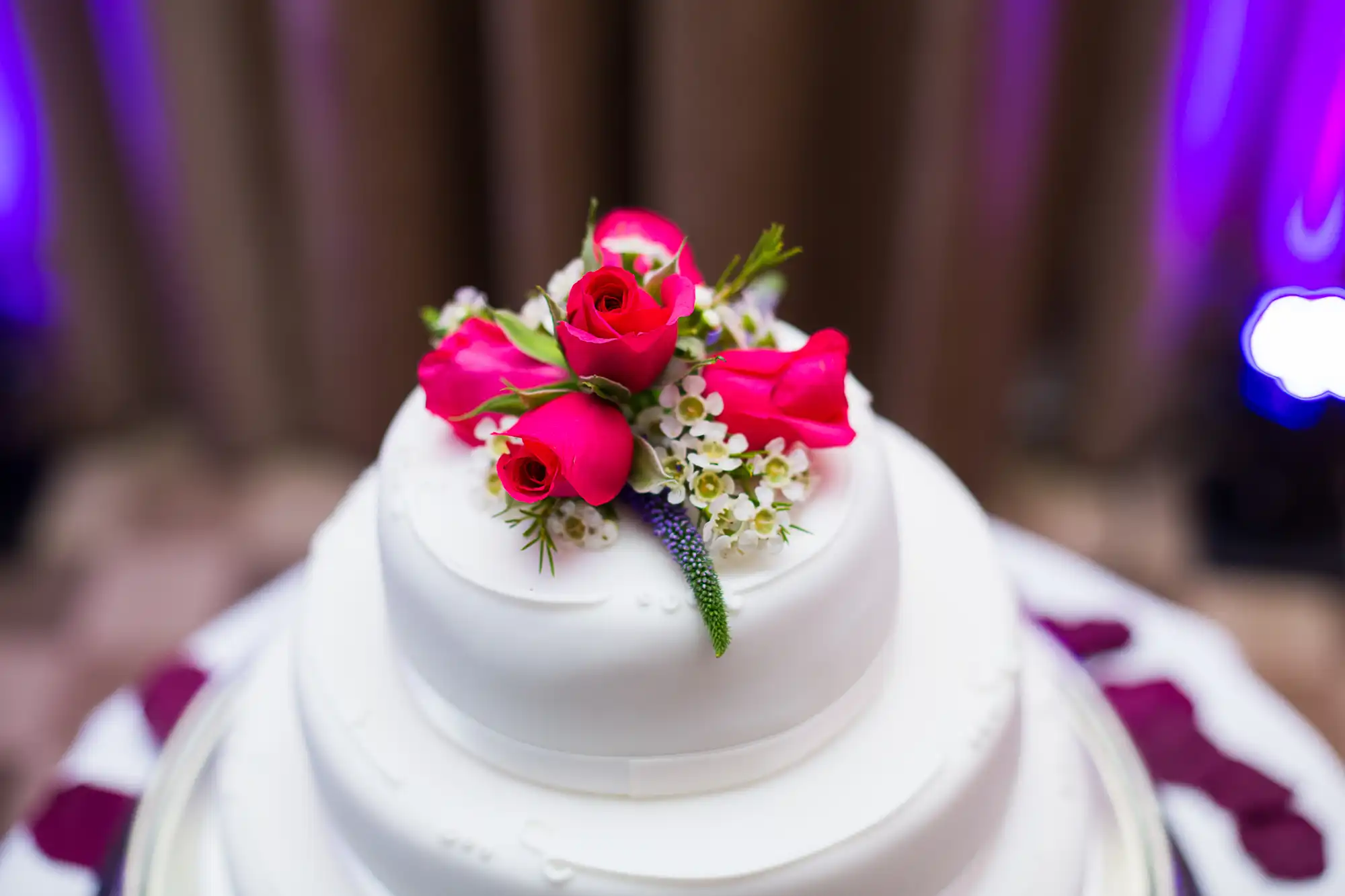 White wedding cake topped with vibrant pink roses and small white flowers, set against a blurred background with purple lighting.