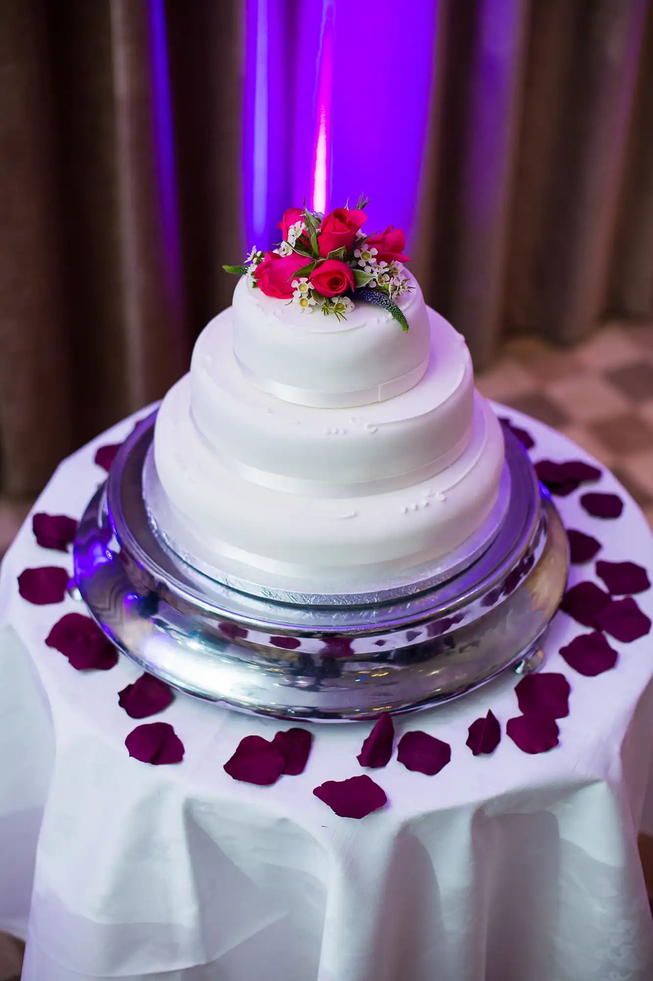 A three-tiered wedding cake with white icing, decorated with red flowers, displayed on a silver stand surrounded by rose petals, lit by a purple light.
