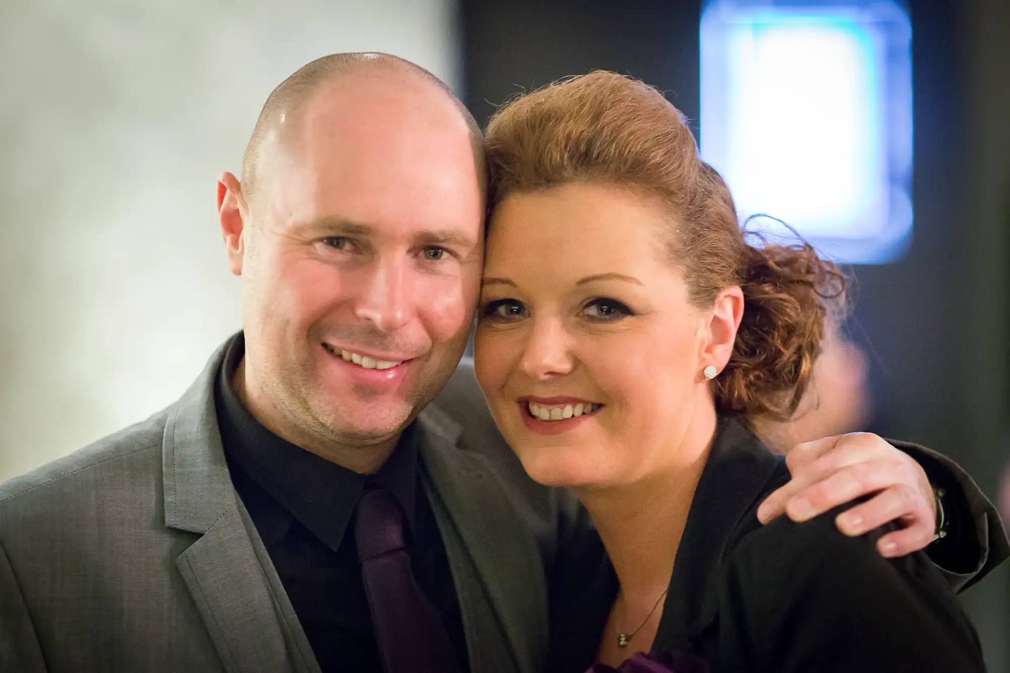 A smiling bald man and a woman with styled brown hair, both dressed in formal attire, posing closely for a photo in a softly lit indoor setting.