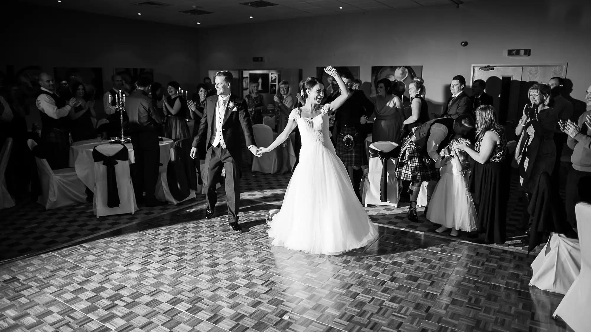 A bride and groom joyously perform their first dance in a ballroom surrounded by applauding guests, capturing a moment of celebration in black and white.