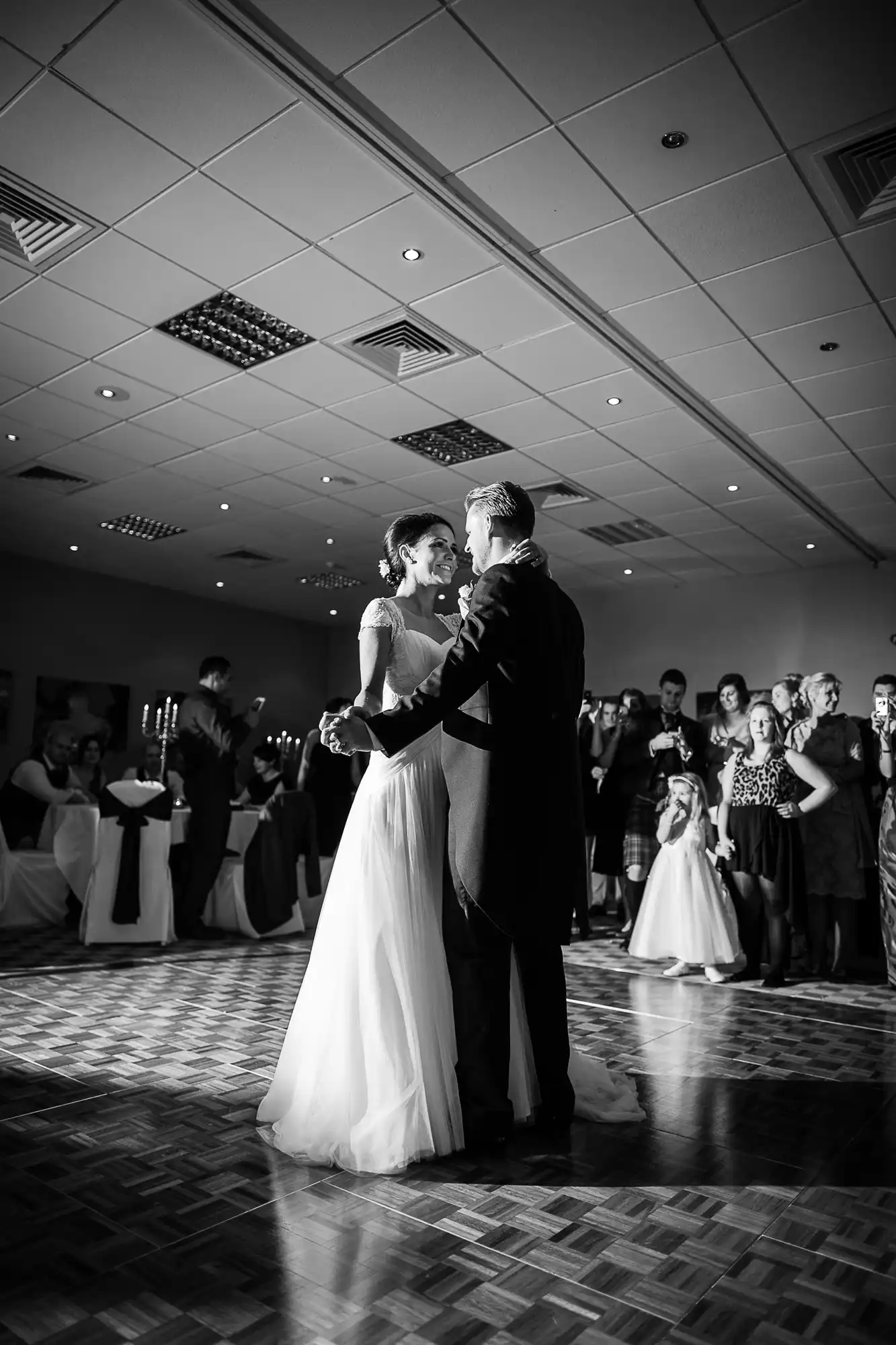 A bride and groom share a dance in a ballroom surrounded by guests, illuminated by overhead lights, captured in black and white.