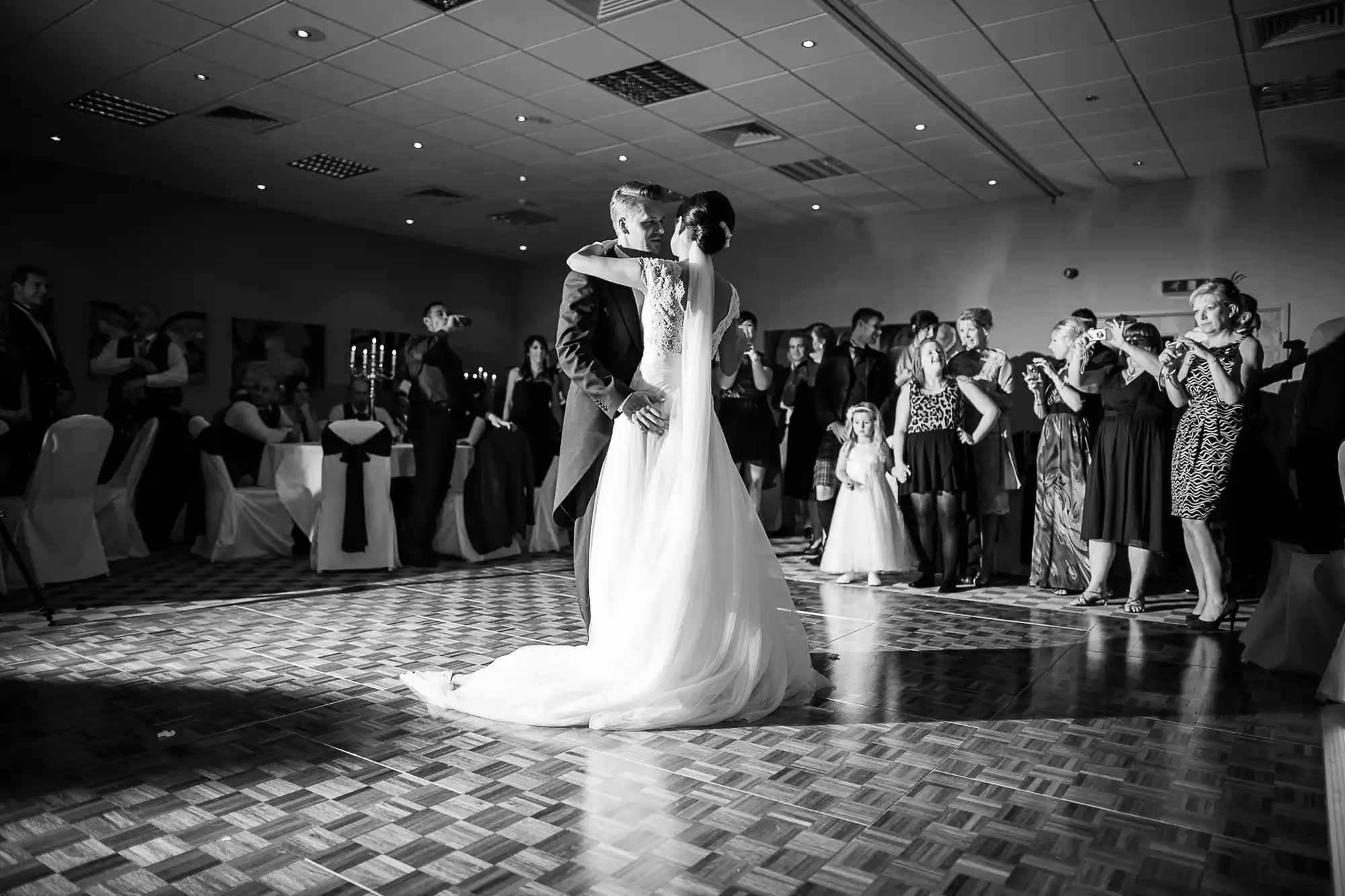 A bride and groom share a kiss on the dance floor as wedding guests look on and applaud in a warmly lit room.