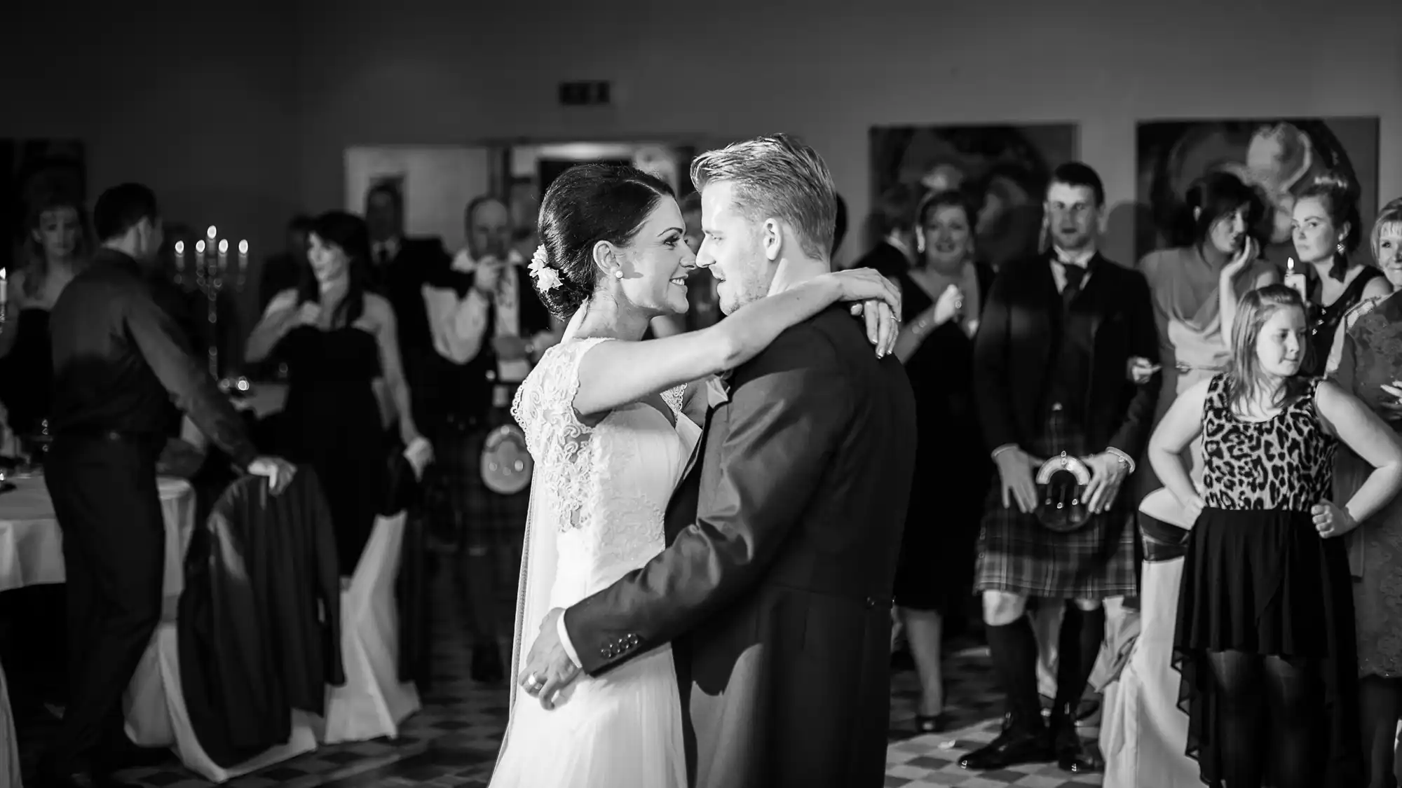 A bride and groom share a dance at their wedding reception, surrounded by guests, in a black and white photo.