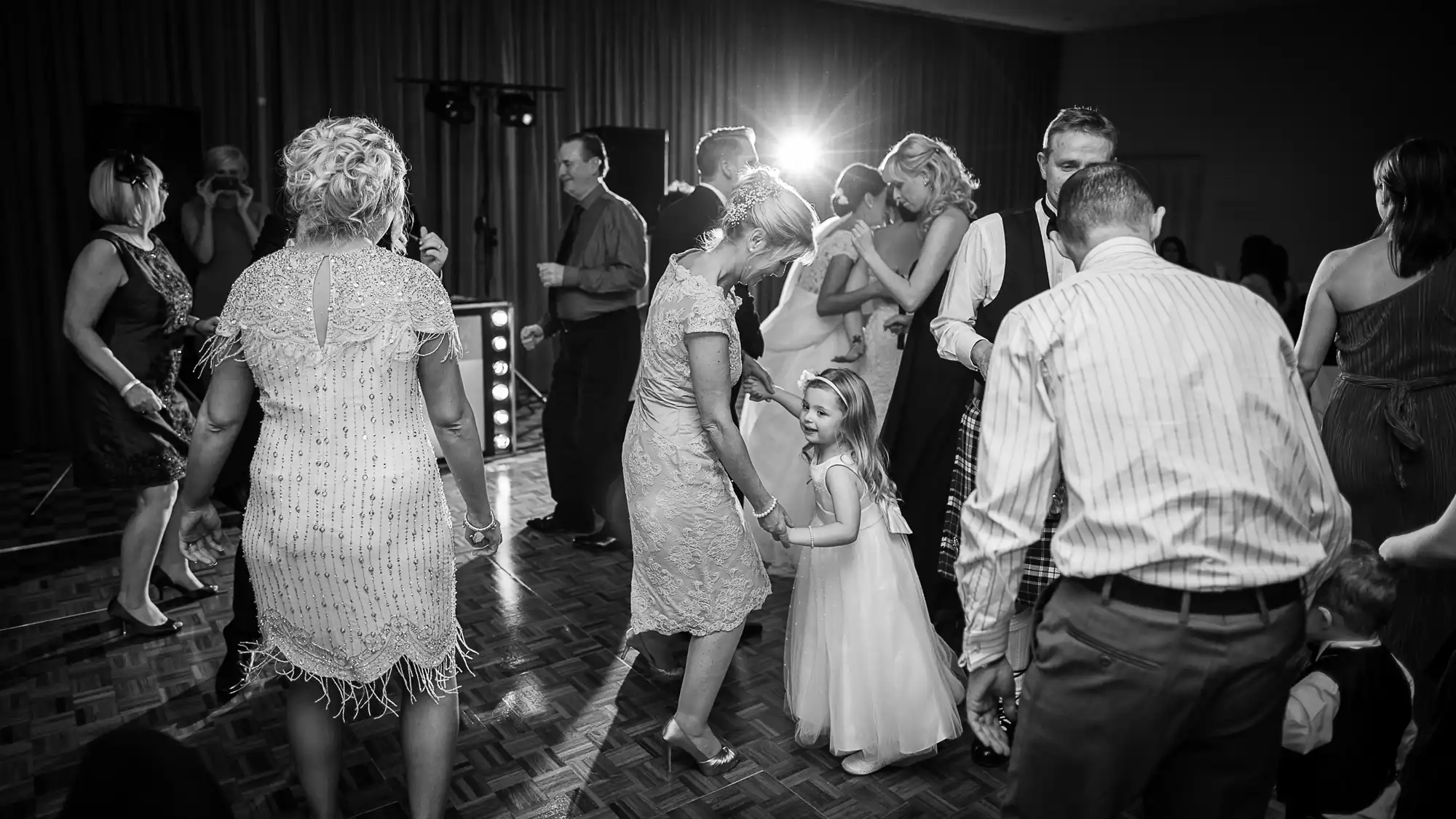Black and white image of people dancing and socializing at an indoor party, with a little girl interacting with adults under a spotlight.