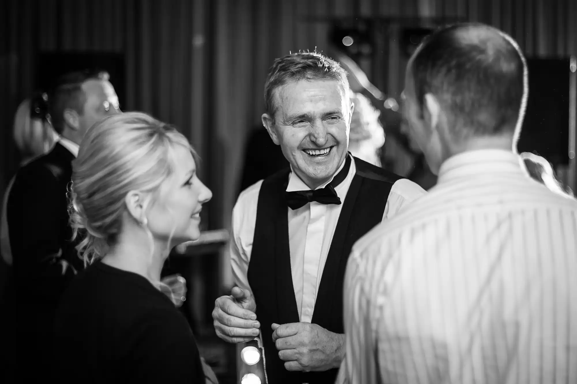 An older man in a tuxedo smiles while chatting with people at a formal event, captured in black and white.
