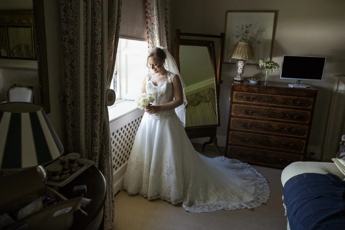A bride in a white gown looks at her bouquet in a vintage-styled room, with elegant furnishings and soft lighting.