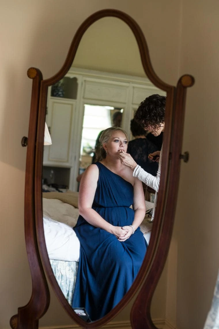 A woman in a blue dress is reflected in a full-length mirror while a makeup artist applies her makeup in a softly lit room.