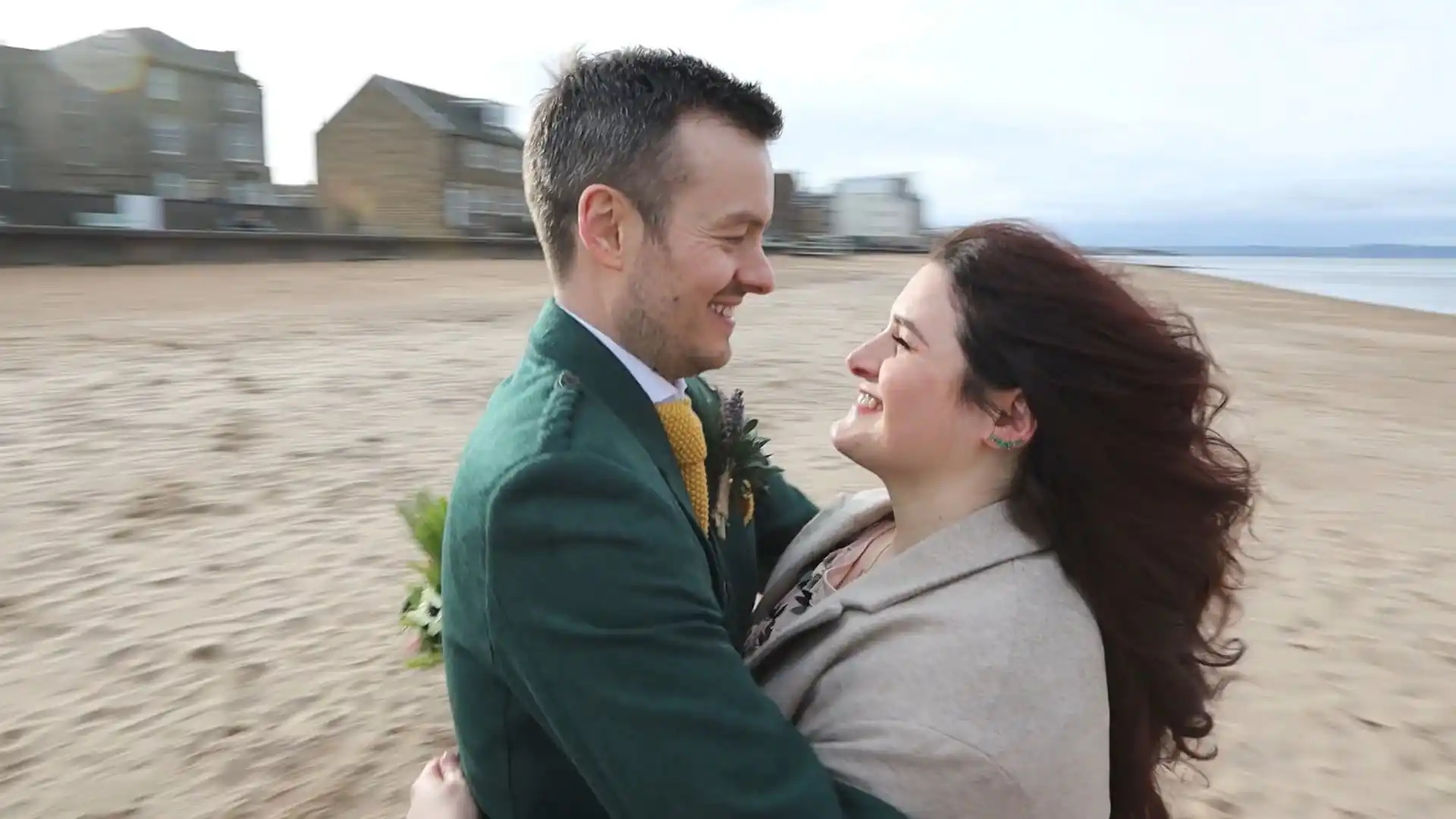 A man and woman embrace and smile at each other on a sandy beach with buildings in the background.