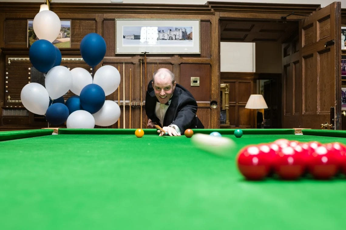 Man aiming a cue at billiard balls on a green table, with blue and white balloons in the background.