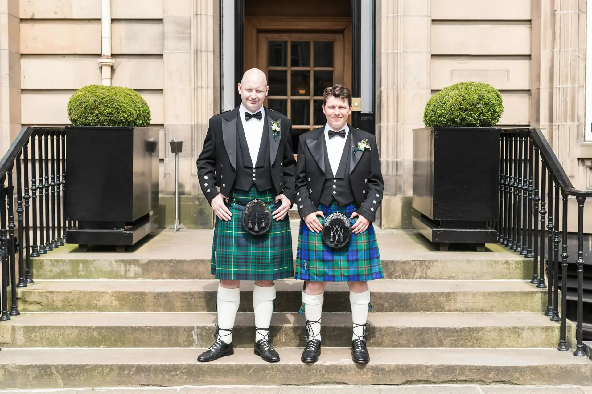 Two men in traditional scottish kilts and jackets standing on steps outside a building, smiling at the camera.
