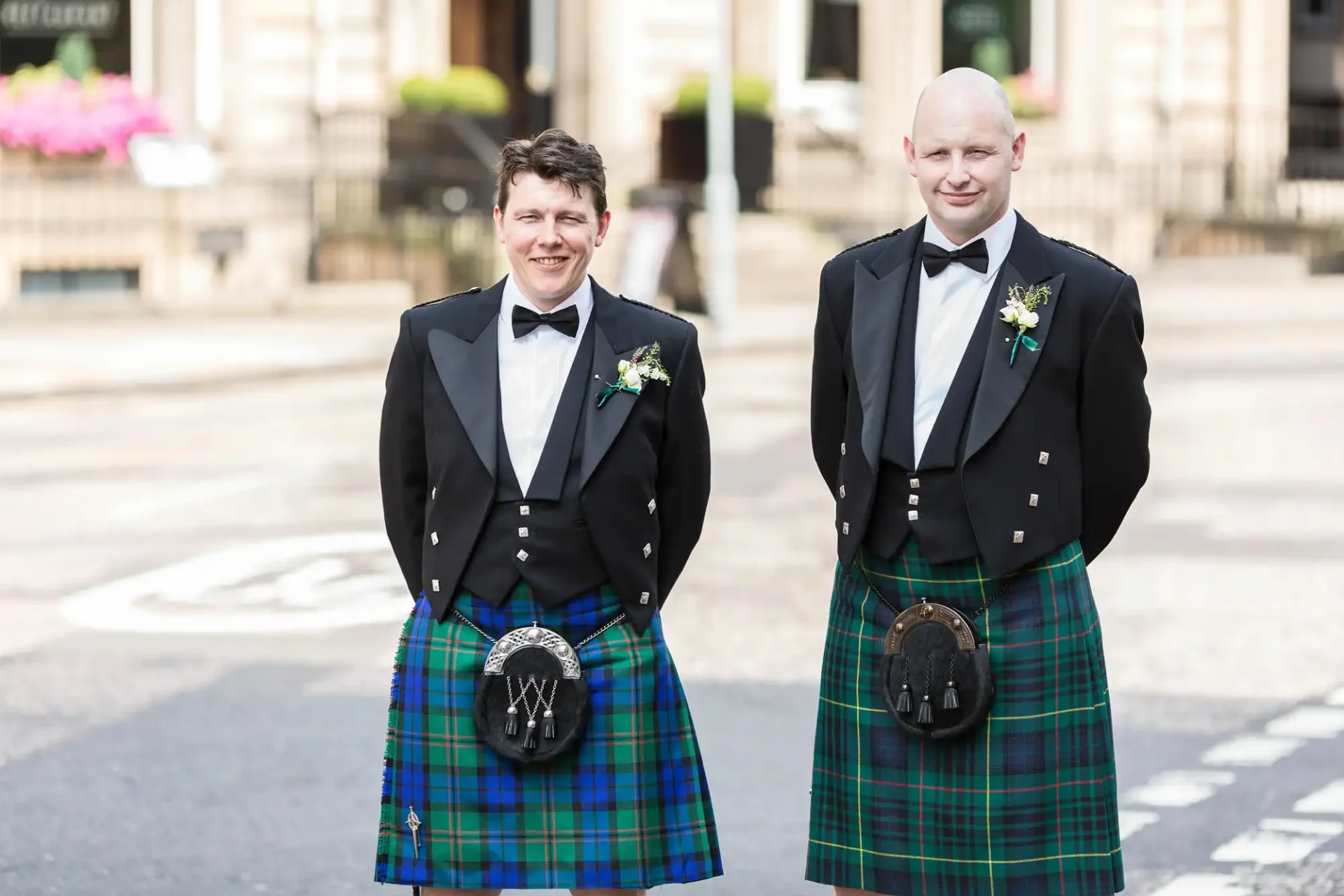 Two men in formal attire with kilts and sporran, smiling on a street, wearing bow ties and boutonnieres.