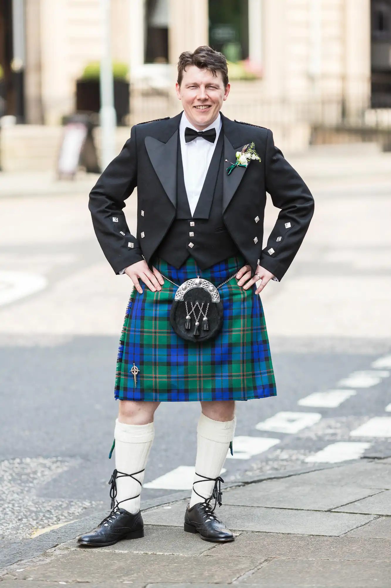 Man in traditional scottish attire including a kilt, black jacket, and sporran, smiling on a city street.