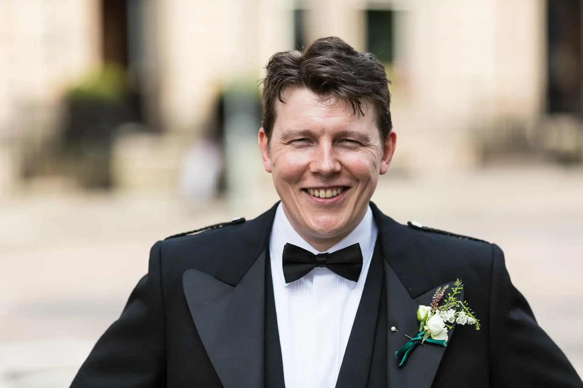 A smiling man in a black tuxedo and bow tie with a boutonniere on his lapel, standing outdoors.