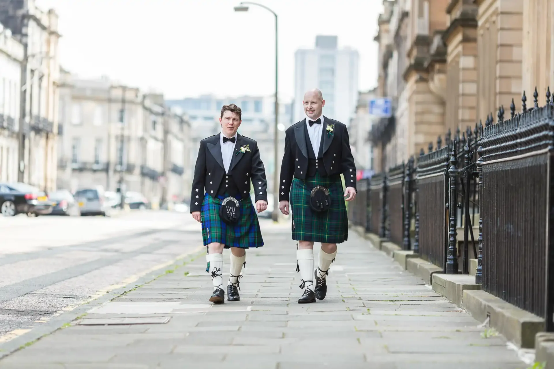Two men dressed in traditional scottish kilts and jackets walking along a city street, with buildings and parked cars in the background.