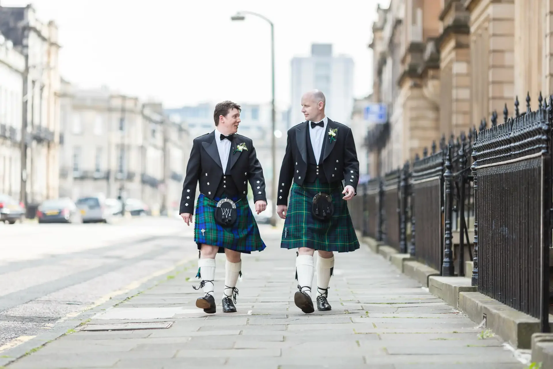Two men in formal kilts and jackets walking on a city street, smiling and conversing.