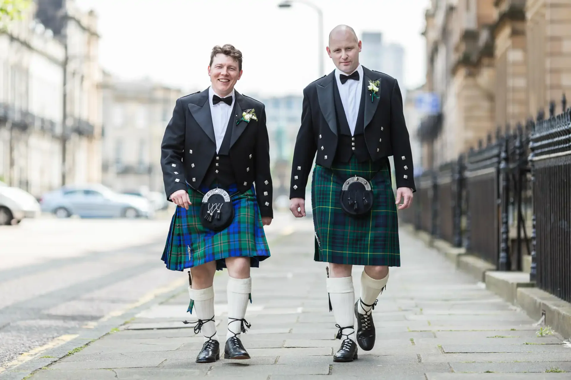 Two men dressed in traditional scottish kilts and jackets, smiling as they walk down a city street.