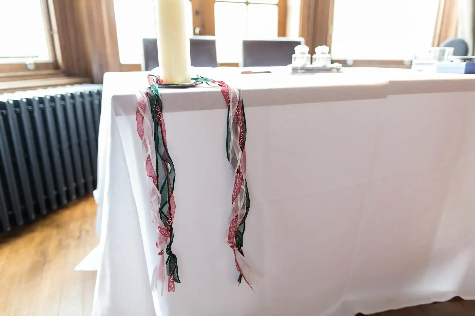 A white tablecloth covers a long table with a decorative ribbon tied to one corner, and items like a candle and stationery set on top, in a room with wooden floors and large windows.