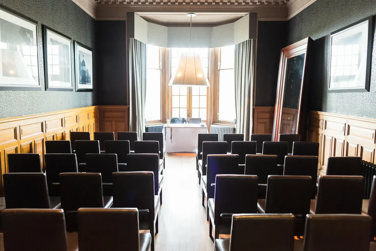 A lecture hall with rows of black chairs facing a podium under a large window, surrounded by wood paneling and grey walls.
