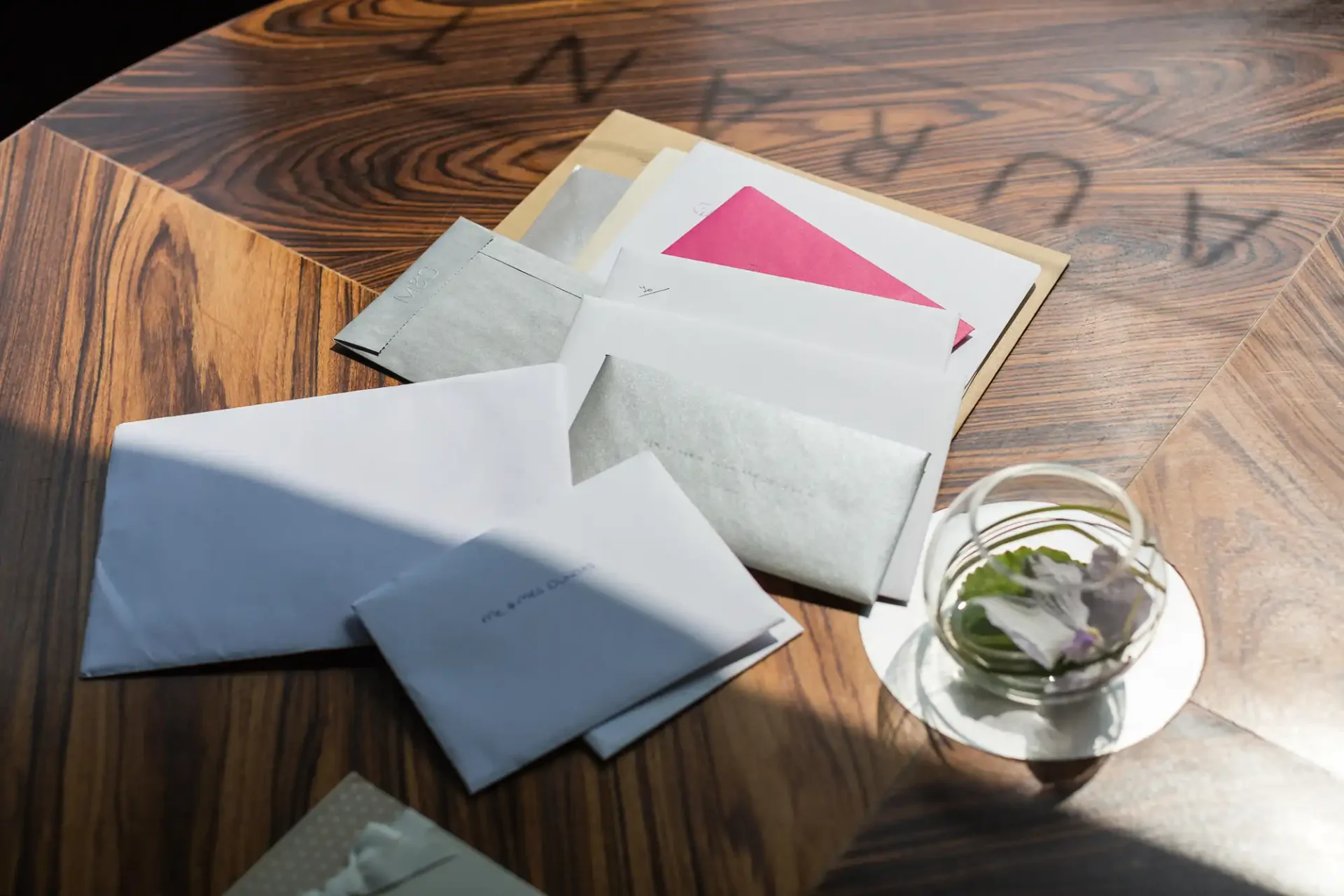 Sunlight shines on a wooden table holding several scattered envelopes and a glass vase with a white flower.