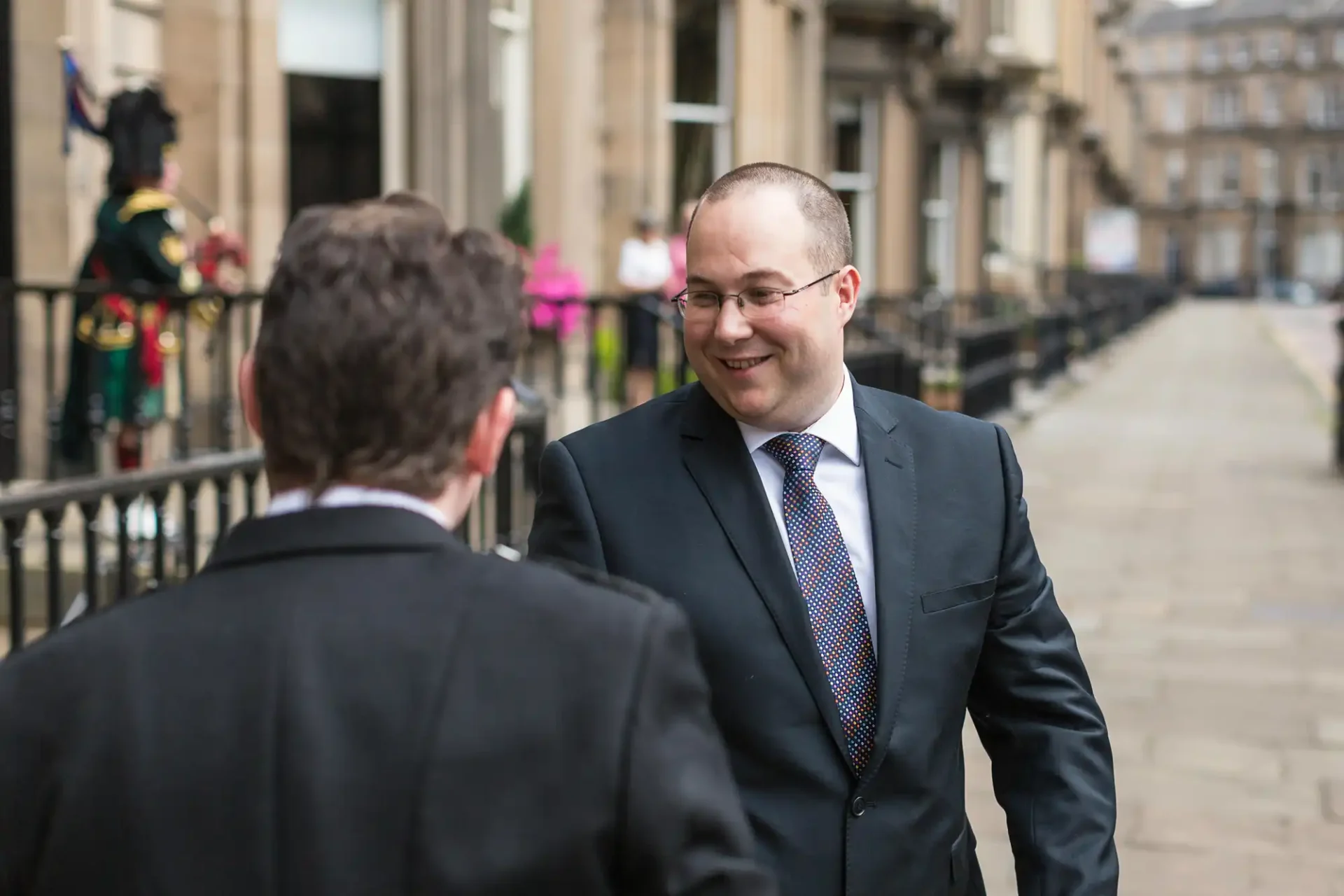 Two men in business suits engaged in a conversation on a city street, with one man facing the camera and smiling.