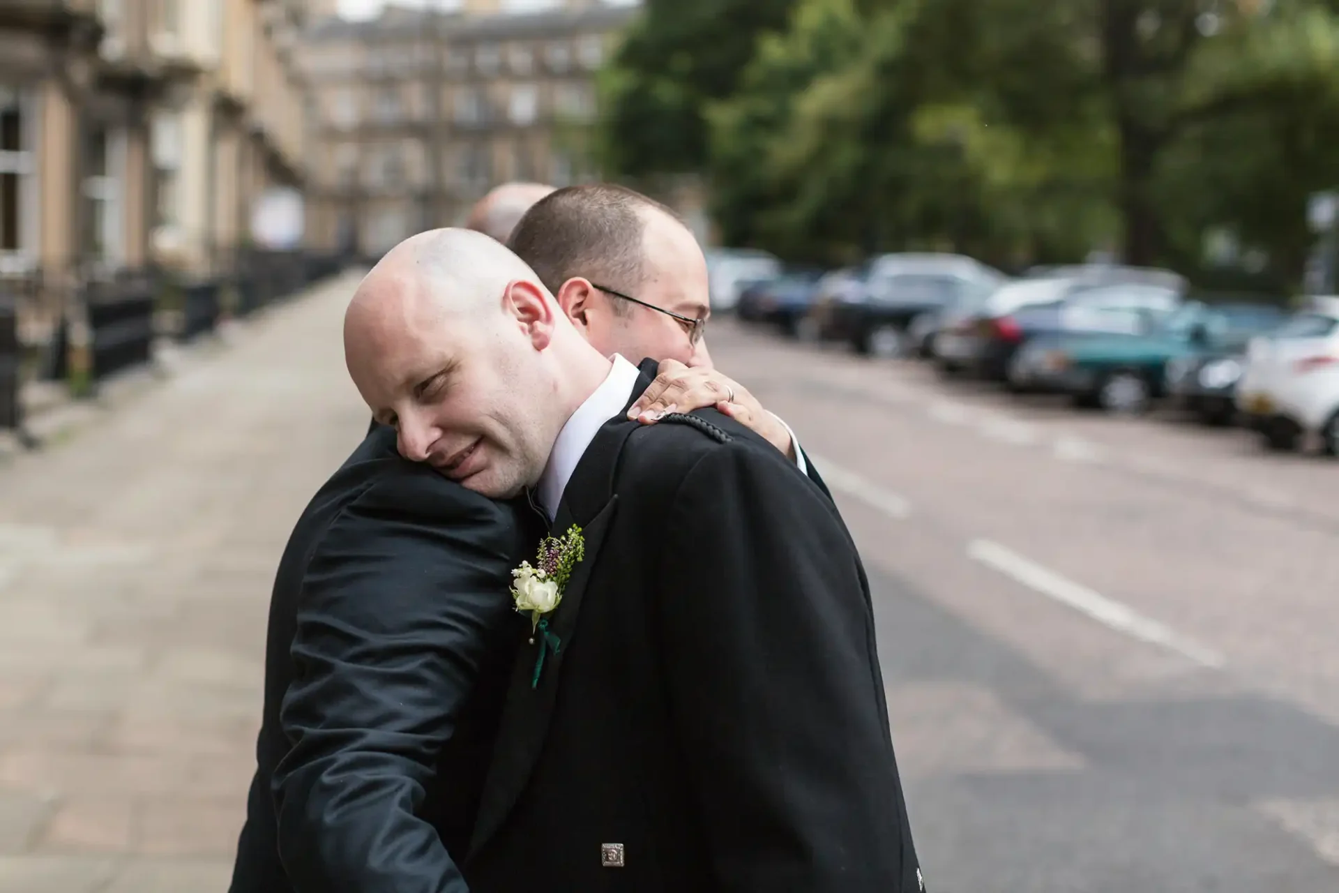 Two men in suits hugging on a street, displaying affection and emotion, with cars and buildings in the background.