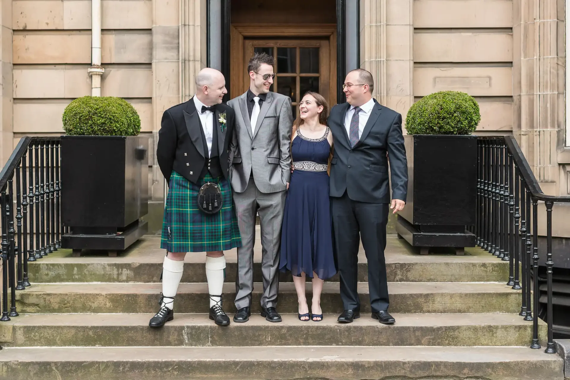 Four people smiling on steps outside a building, one man in traditional scottish kilt, others in formal wear.