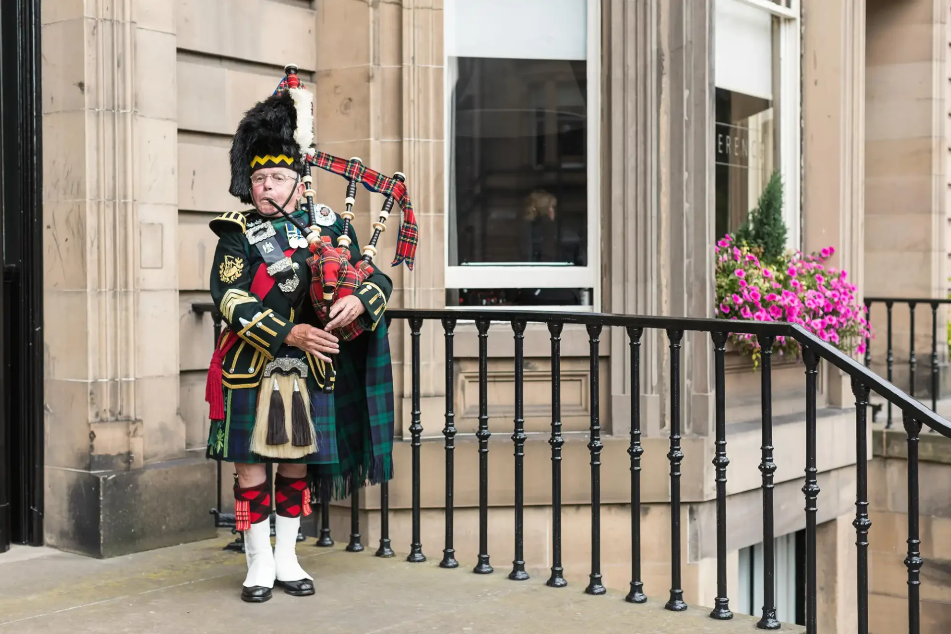 A bagpiper in traditional scottish attire playing outside a building.