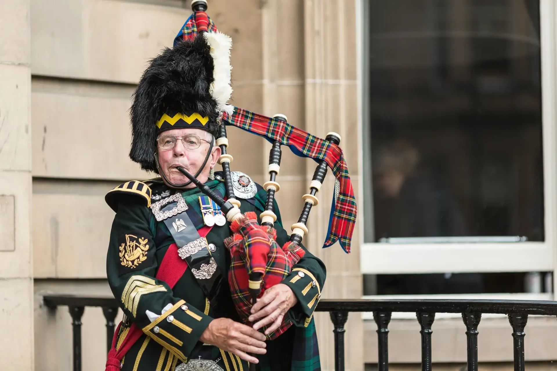 A bagpiper in traditional scottish attire, including a feathered hat and tartan kilt, plays the bagpipes outside a building.