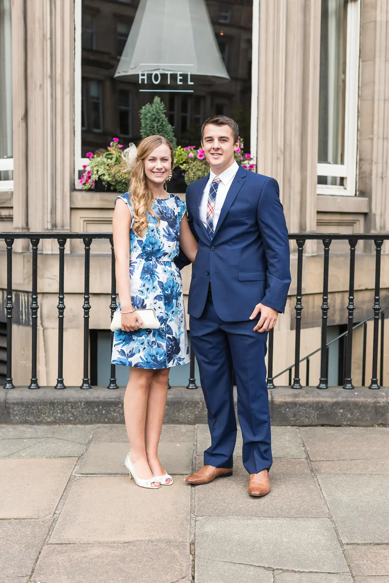 A young couple smiling and standing in front of a hotel, with the woman wearing a floral blue dress and the man in a blue suit.