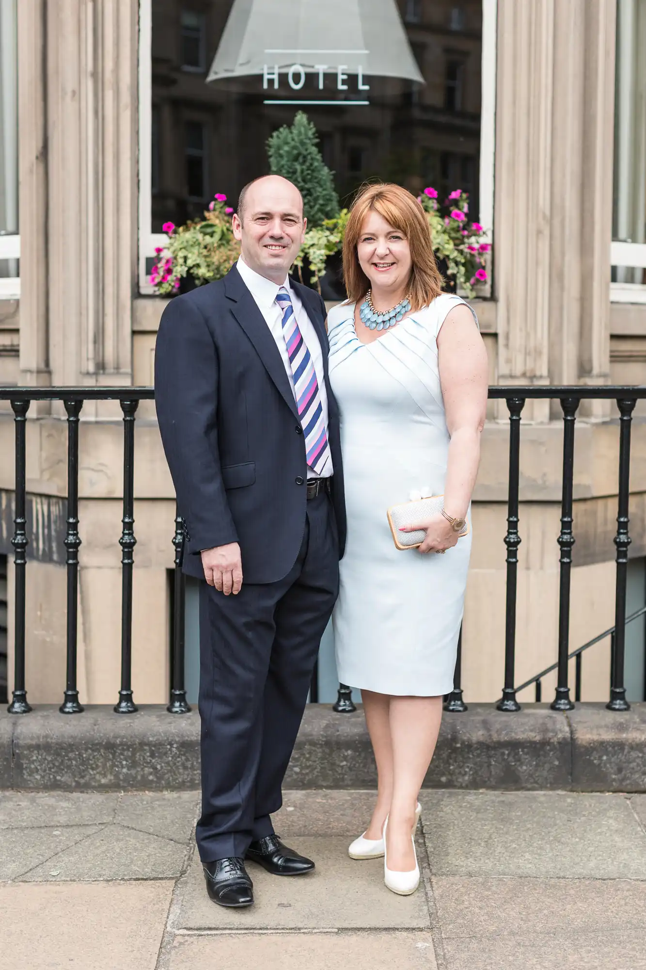 A man and a woman dressed in formal attire standing in front of a hotel entrance with floral decorations.