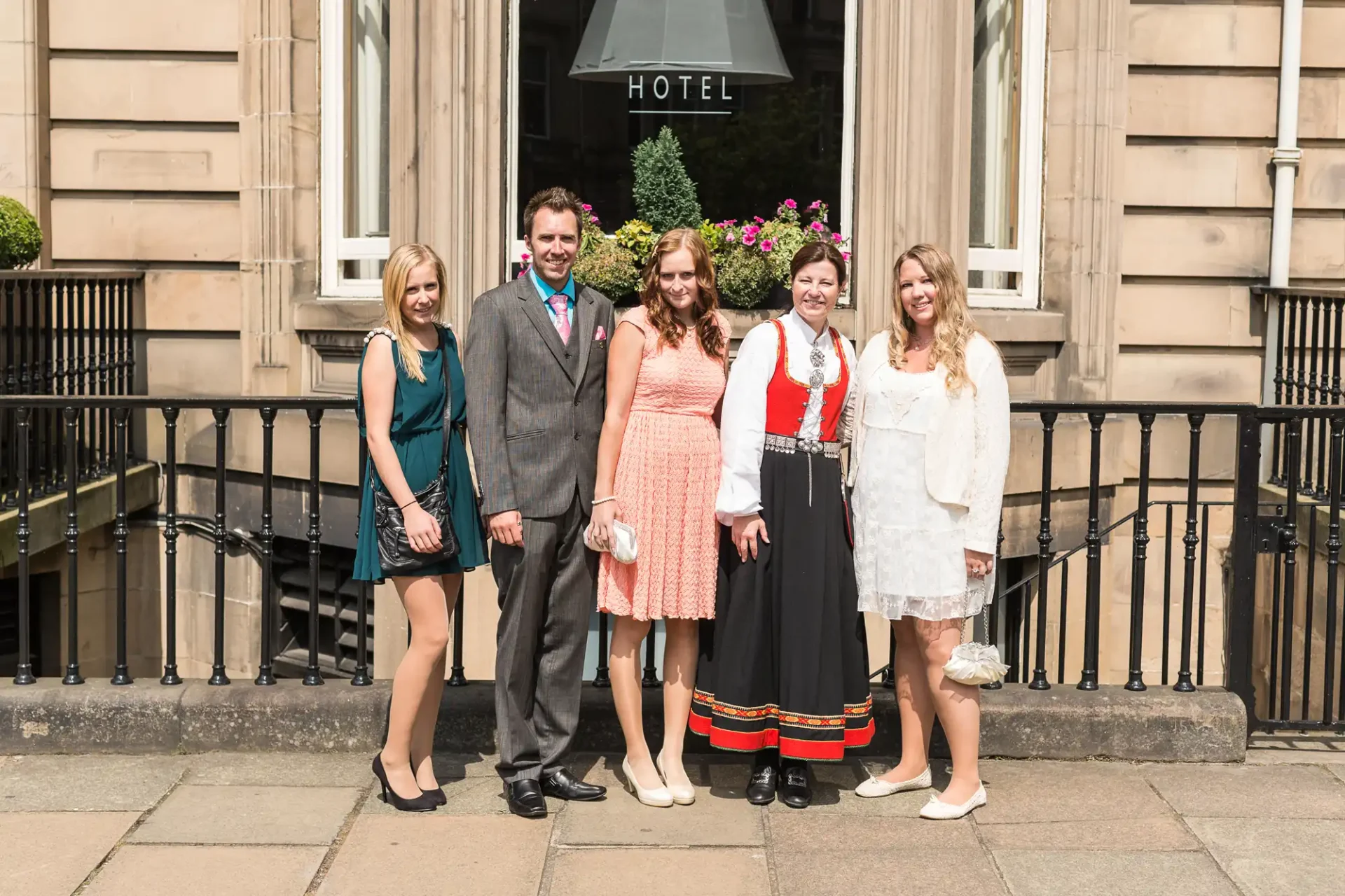 Five people posing in front of a hotel entrance, two women in traditional scandinavian dresses, one man in a suit, and two women in modern dresses.