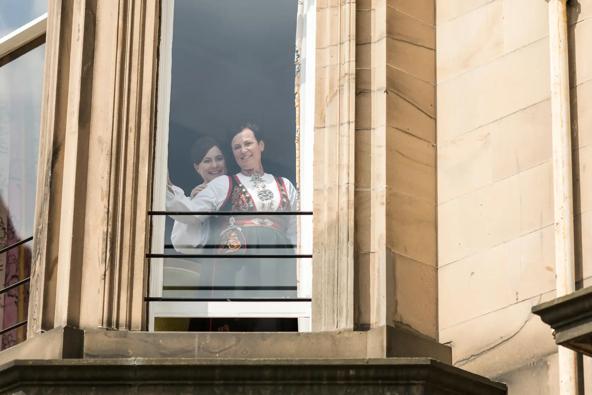 Two women in traditional embroidered dresses smiling and standing behind a balcony railing, viewed from outside a building.