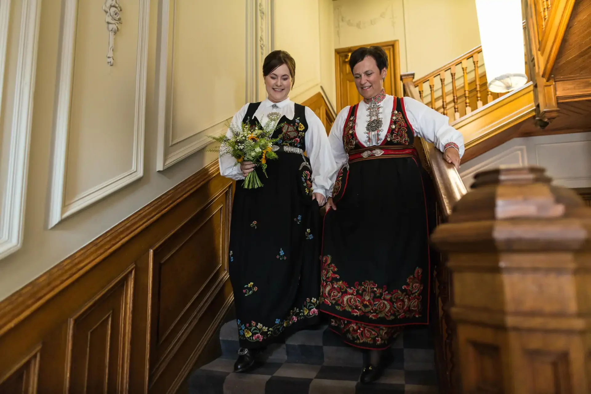 Two women in traditional norwegian dresses descending a staircase, smiling and holding hands, one carrying a bouquet.