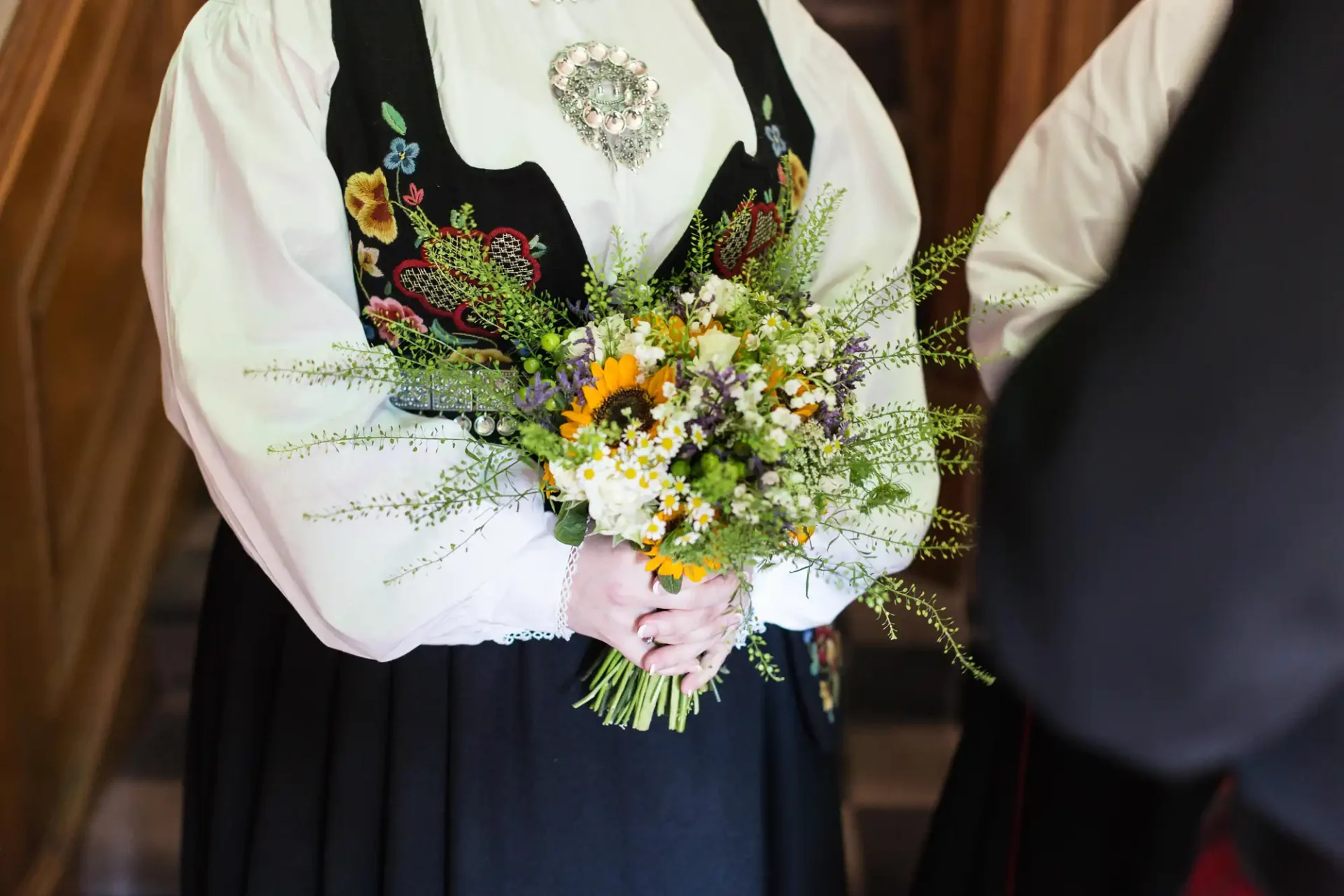 A person in traditional dress holding a bouquet of wildflowers, focusing on the hands and flowers, with another person partly visible.